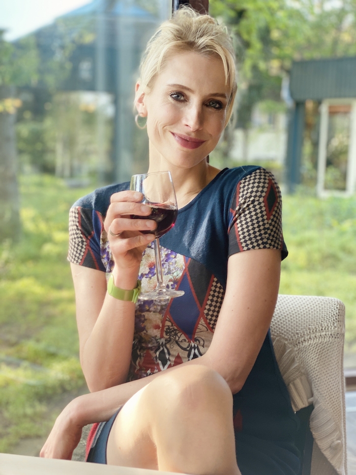 Elischeba Wilde im blauen Kleid mit einem Glas Rotwein in der Hand